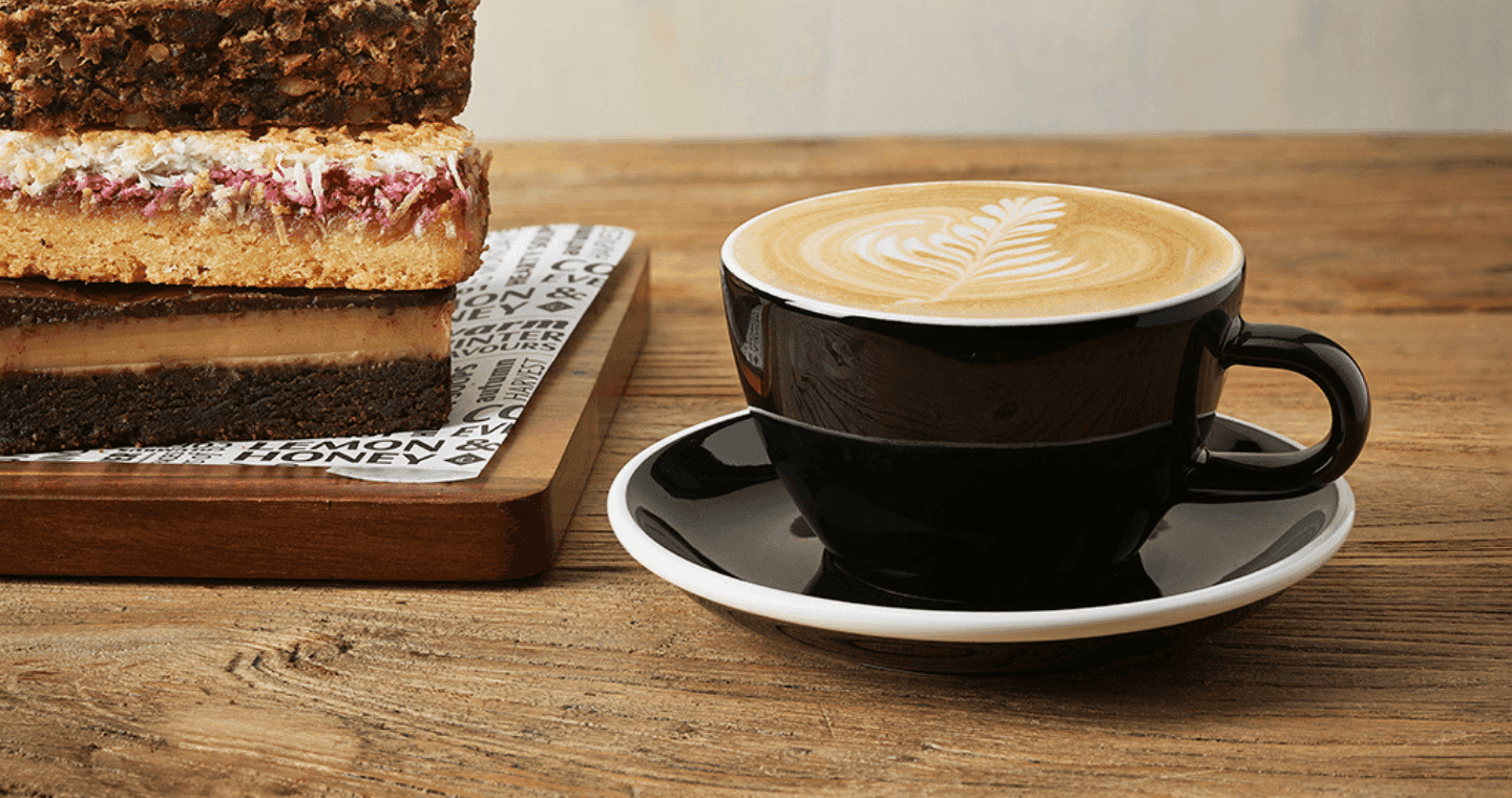 A black coffee cup with a flat white inside and three slices stacked beside on a board
